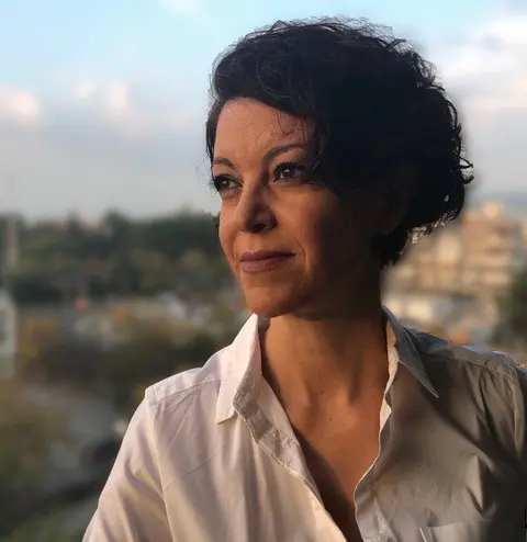 Iphigenia Taxopoulou looking to the side of the frame in a white shirt, with Greek nature backdrop